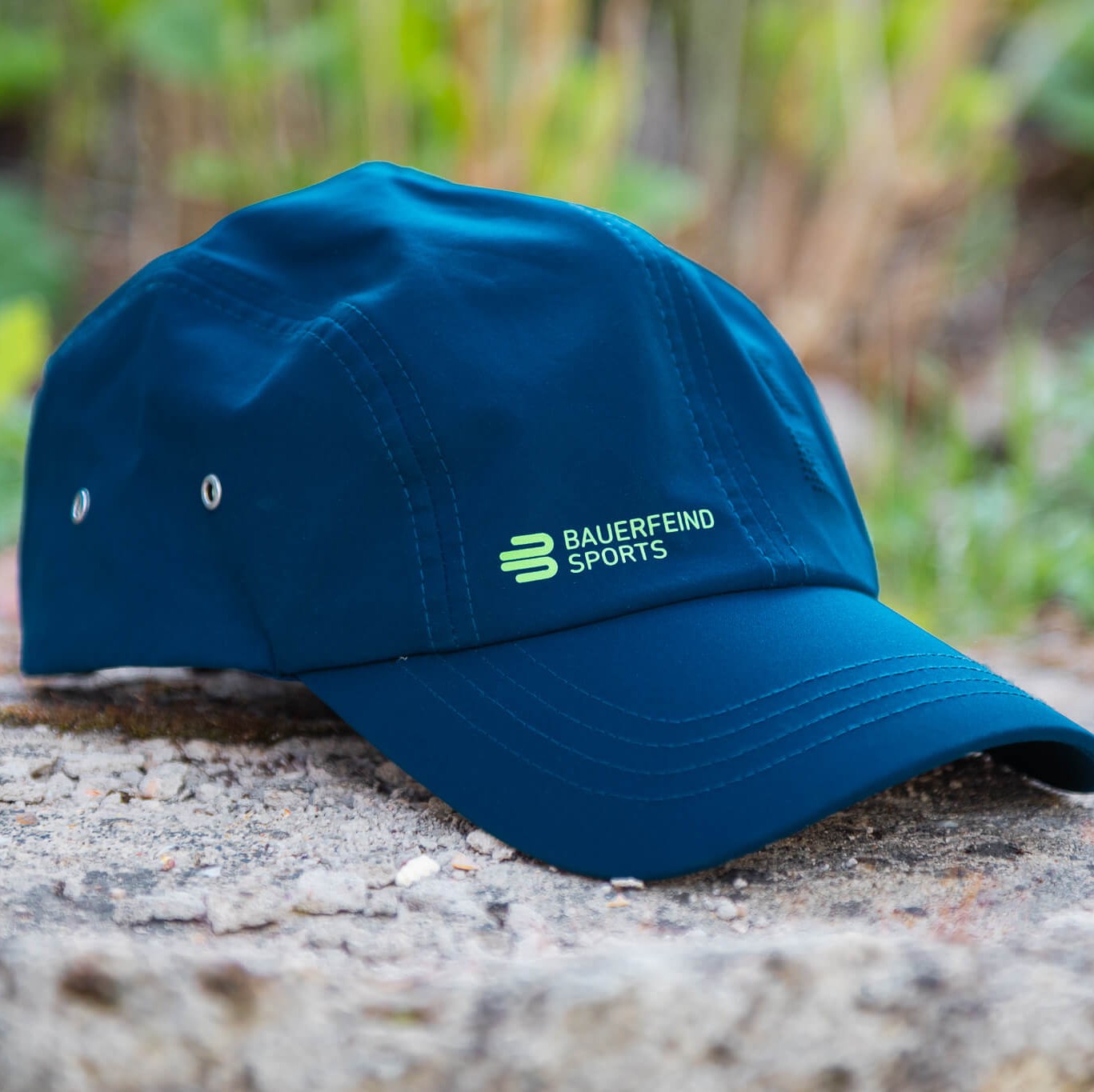 Blaue Bauerfeind Sports Cap mit grünem Logo liegt auf einem Stein im Outdoor-Bereich, umgeben von unscharfem Grün im Hintergrund – ein sportlicher Begleiter für aktive Tage draußen.