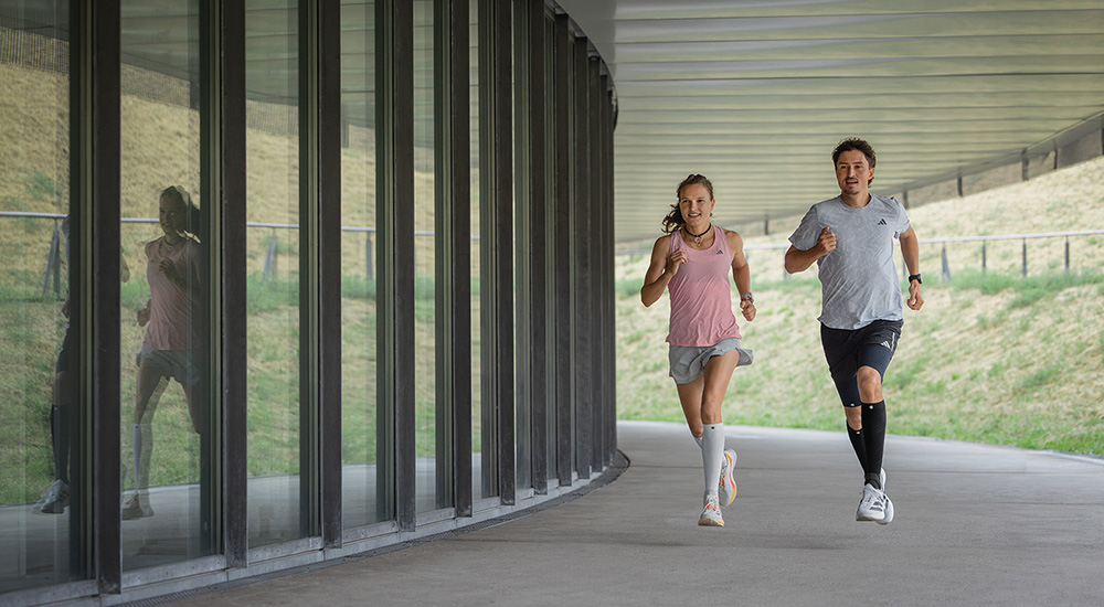 Gemeinsam stark – Kilometer für Kilometer. Zwei Läufer – eine Frau und ein Mann – joggen gemeinsam unter einer überdachten Passage. Die Frau trägt weiße Compression Sleeves, der Mann schwarze. Beide bewegen sich synchron und wirken konzentriert und energiegeladen.